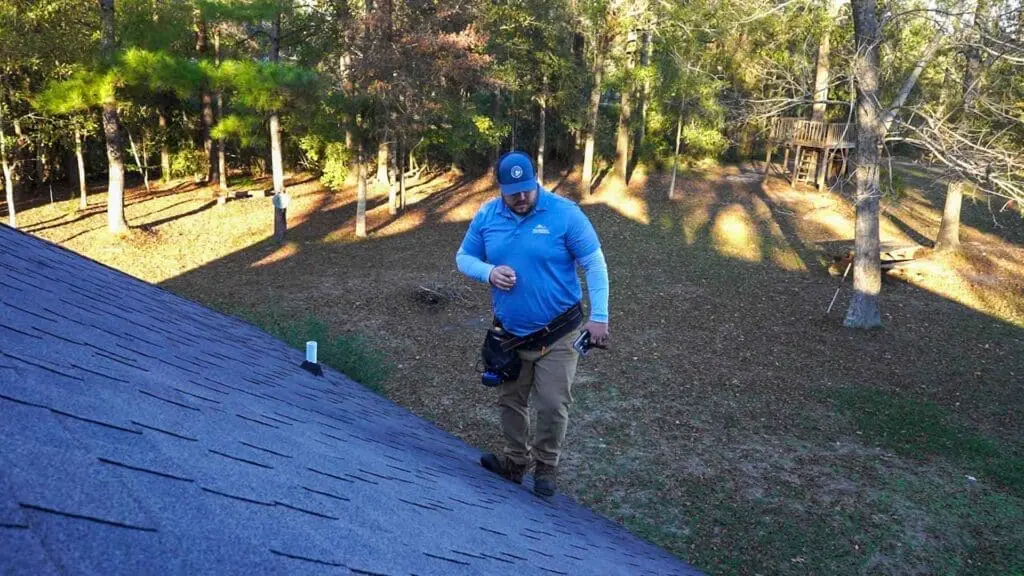 A Wise Builders Roofing and Renovations project manager on a ladder inspecting a Gulf Coast roof with a tablet during a free roof inspection, while the homeowner watches from the driveway.